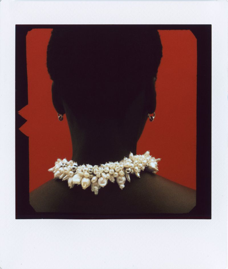 Model viewed from behind wearing an ornate clustered pearl necklace against a bold red backdrop in a classic polaroid film frame.