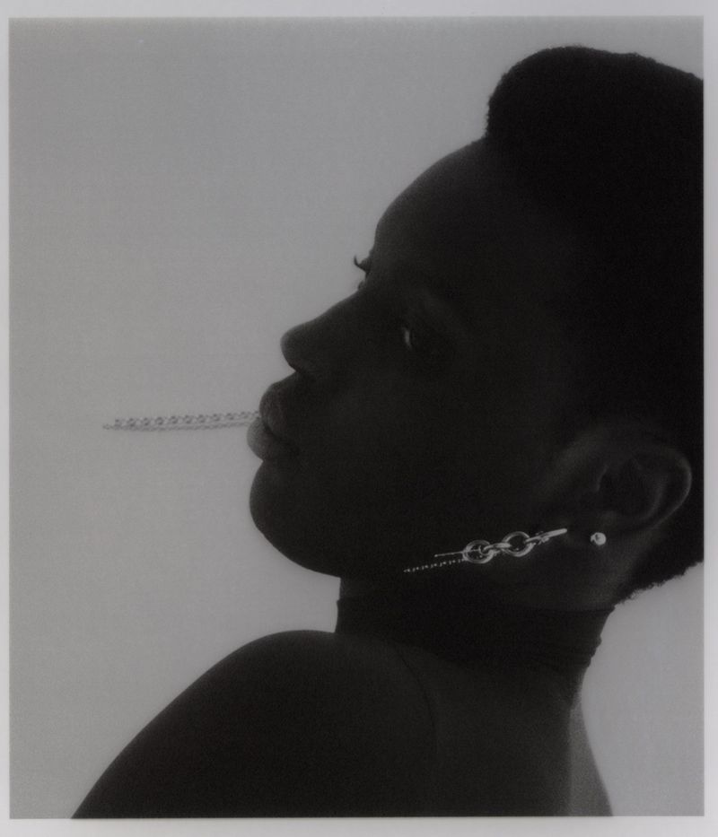 Monochromatic studio shot of a black woman in profile with modern chain earrings and a measuring stick as a prop in her mouth.