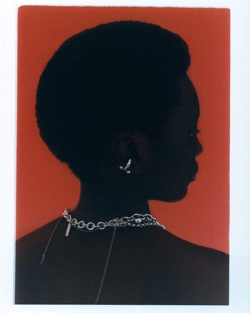 Profile of an adult female with dark skin, wearing a silver chain necklace and earrings against an orange background.