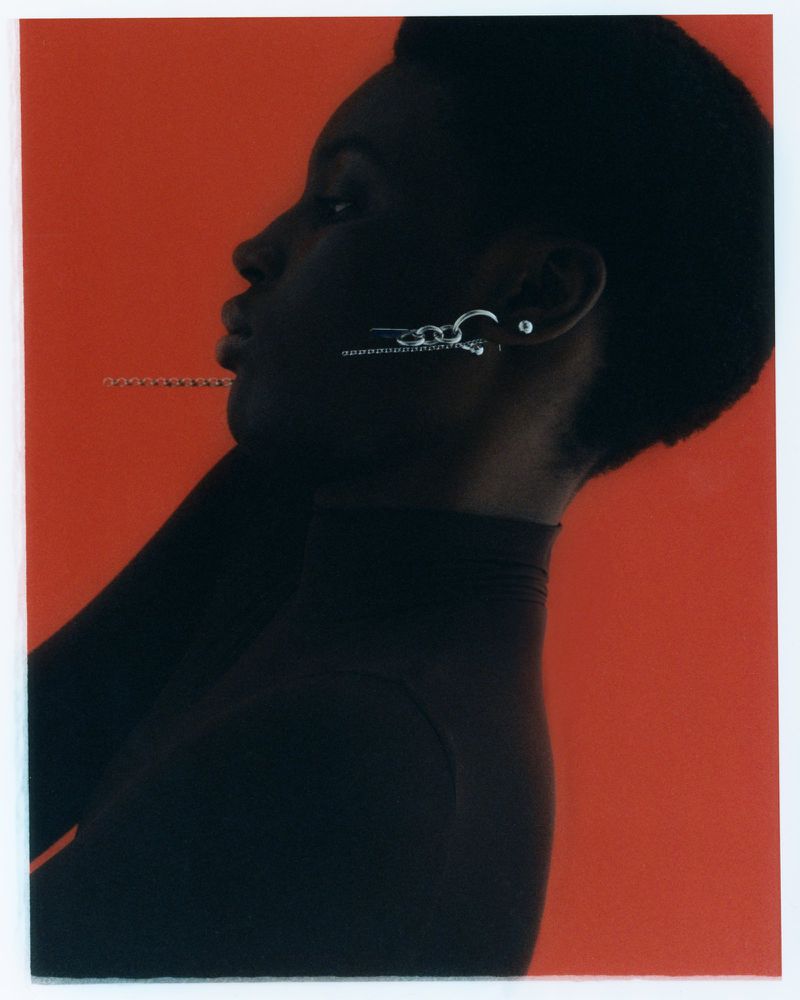 Profile of an adult female model with dark skin in a black turtleneck, wearing silver earrings against an orange-red gradient background.