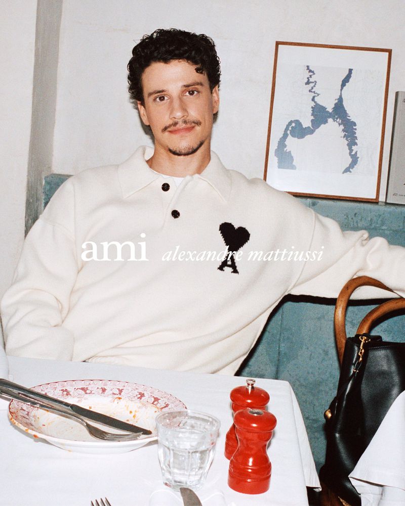 Indoor portrait of an adult male at a restaurant table wearing a cream-colored Ami sweater with a large black heart logo.