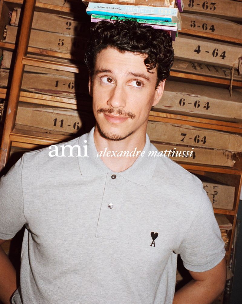 Adult man with books on his head smiles in a close-up studio shot, wearing a grey polo shirt, standing against a wood paneled background.