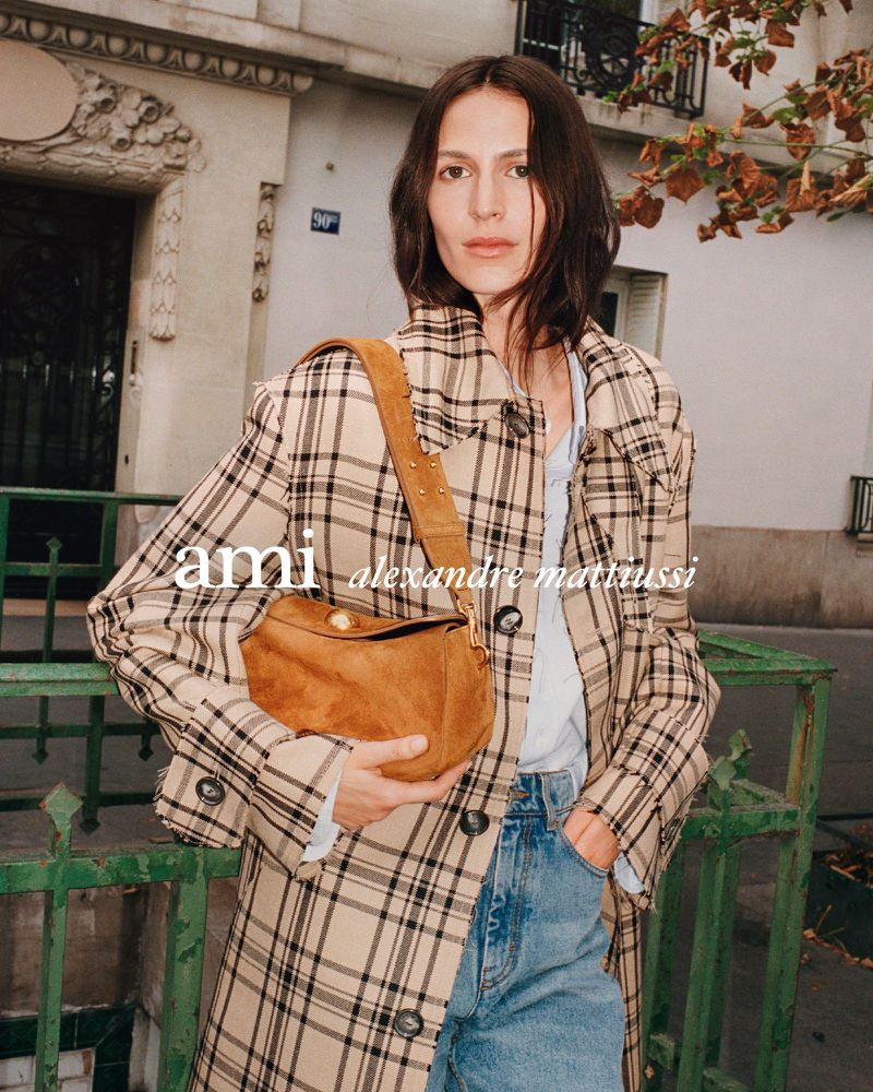 An adult female stands outside wearing a plaid coat, jeans, and holding a brown suede bag, photographed at eye-level with natural lighting.