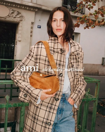 An adult female stands outside wearing a plaid coat, jeans, and holding a brown suede bag, photographed at eye-level with natural lighting.