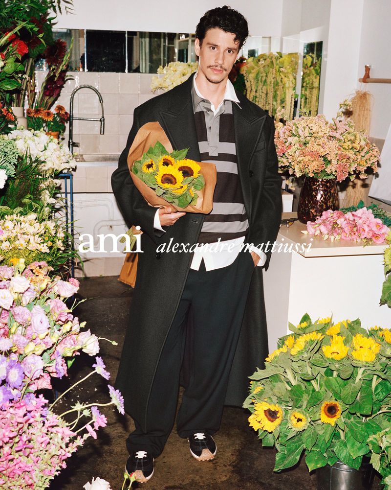 Man in coat stands in flower shop holding sunflowers. He wears a striped shirt and dark pants. Bouquets and flowers surround him.