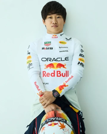 Formula 1 driver Yuki Tsunoda wearing a white Red Bull racing suit and black Tag Heuer watch while holding a helmet.