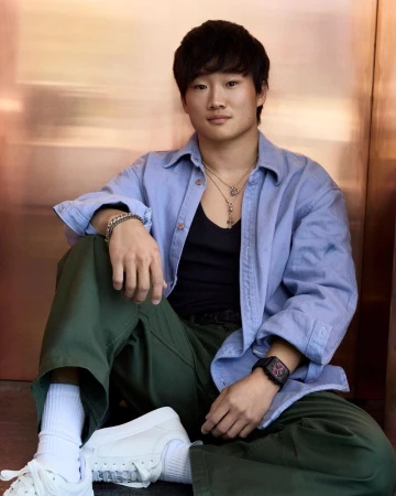 Yuki Tsunoda seated in an open blue shirt, dark tank top, olive trousers, and white sneakers against a copper metallic wall.