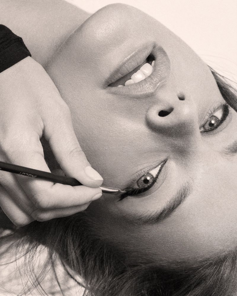 Close-up shot of a model getting her eyeliner applied by a makeup artist with a thin brush in a beauty studio, monochrome.