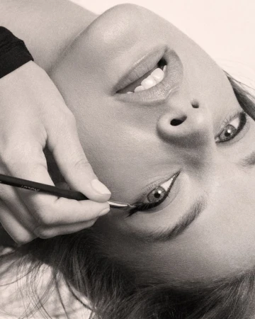Close-up shot of a model getting her eyeliner applied by a makeup artist with a thin brush in a beauty studio, monochrome.
