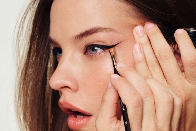 Close-up of a woman applying black eyeliner with a thin brush, her blue eyes and fair skin illuminated in a makeup artistry shot.
