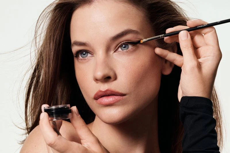 A woman with fair skin is having her eye makeup done with a fine brush and black eyeliner gel in a beauty advertising campaign.