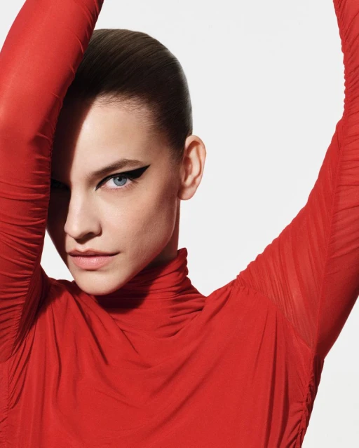 Fashion portrait of a woman with red long-sleeved shirt looking at the camera with eyeliner makeup against a white background.