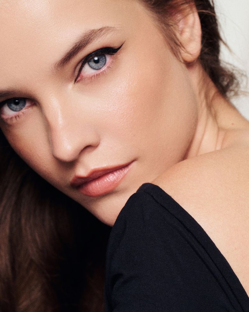 Close-up portrait of a woman with blue eyes and brown hair wearing black eyeliner and a black top against a white background.