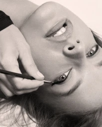Close-up monochrome portrait of model Barbara Palvin receiving black winged eyeliner application with an angled brush.
