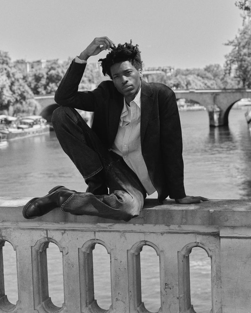 Black and white portrait of a Black man in a jacket and jeans sitting on a ledge with a river and bridge in the background on a sunny day.