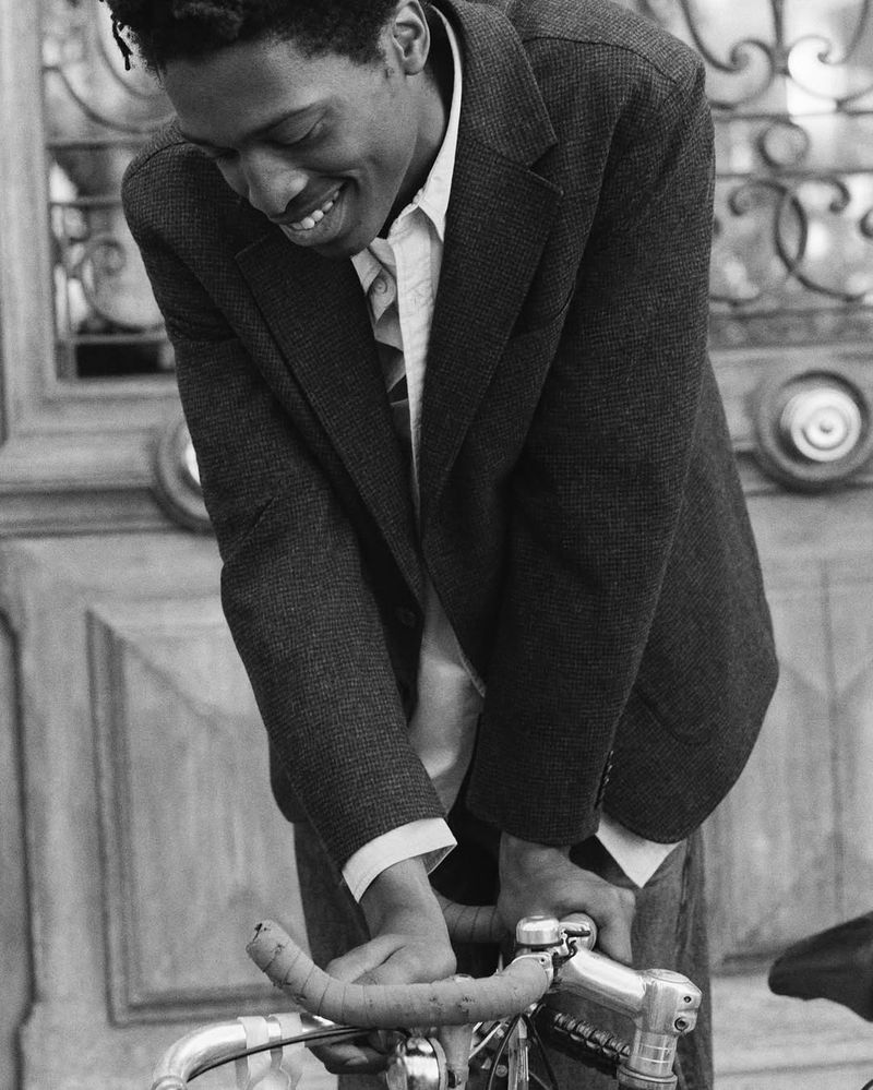 Black and white shot of a smiling man in a blazer with a bicycle on a city street, showcasing classic urban style.