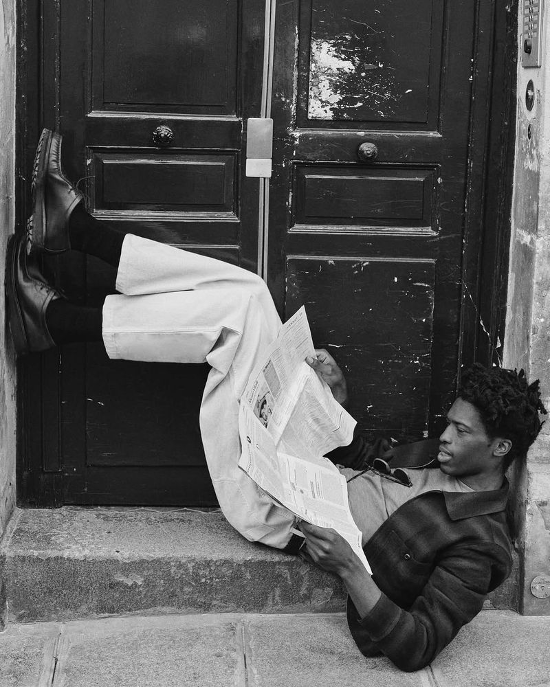 Black and white image of a Black man lying on steps, reading a newspaper, wearing trousers, dress shoes, a jacket, and black socks.