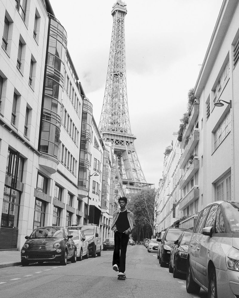 Black and white shot of a man skateboarding in Paris with the Eiffel Tower in the background; urban street scene with cars and buildings.