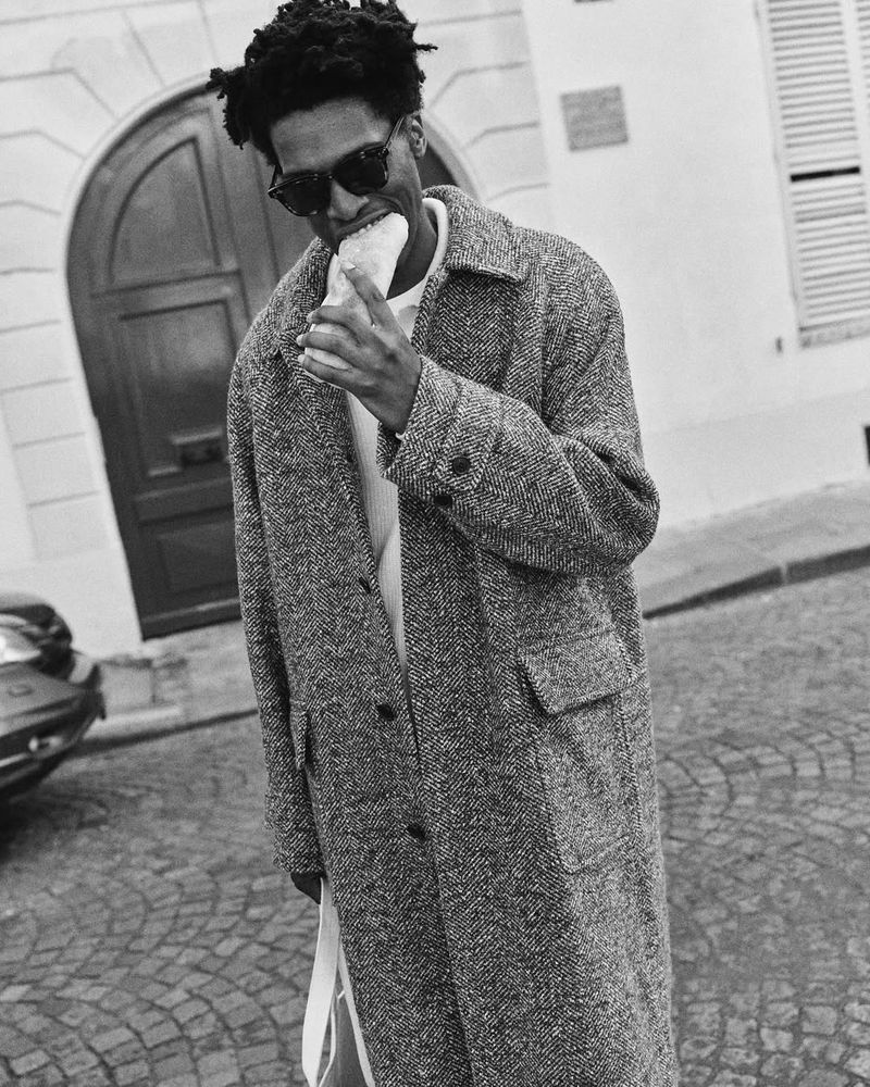 An African-American man in sunglasses and a gray coat eats a baguette on a cobblestone street in front of a building with an arched door.
