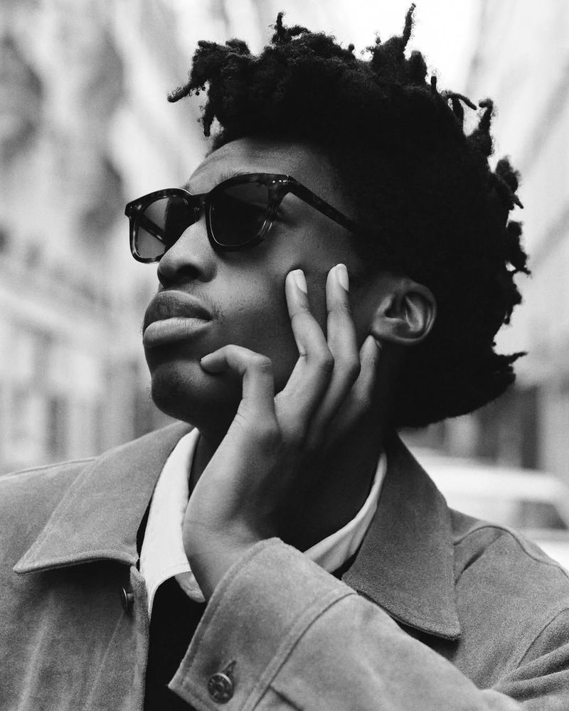 Monochrome portrait of an African-American man wearing sunglasses and a tan jacket in an urban outdoor setting on a sunny day.