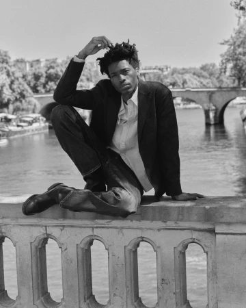 Monochrome portrait of a man squatting on a stone bridge overlooking a river, wearing a dark blazer and jeans.