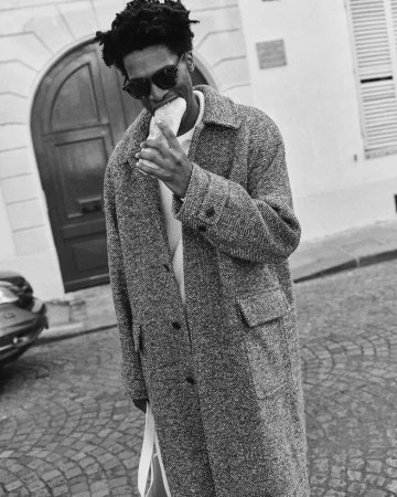 Tamel Lee wearing dark sunglasses and a long tweed overcoat biting into a baguette on a cobblestone street.