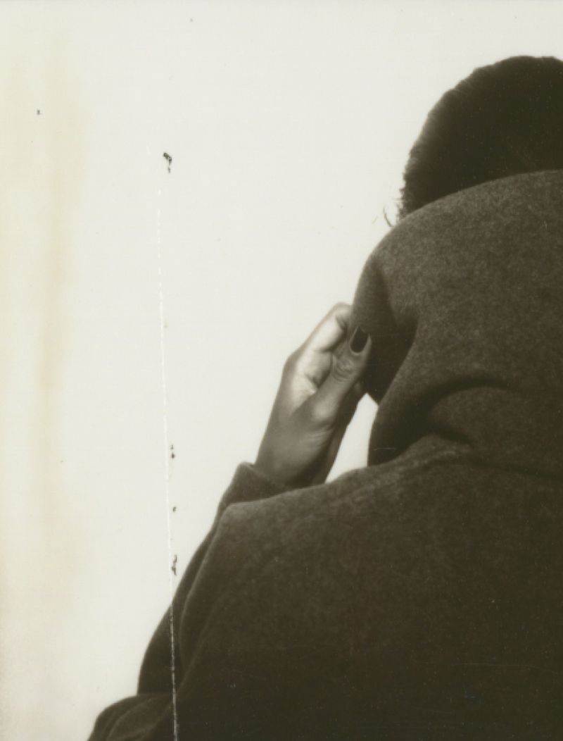 An adult woman in a dark top sits with her hand near her face against a light background, displaying black manicure and dark hair.