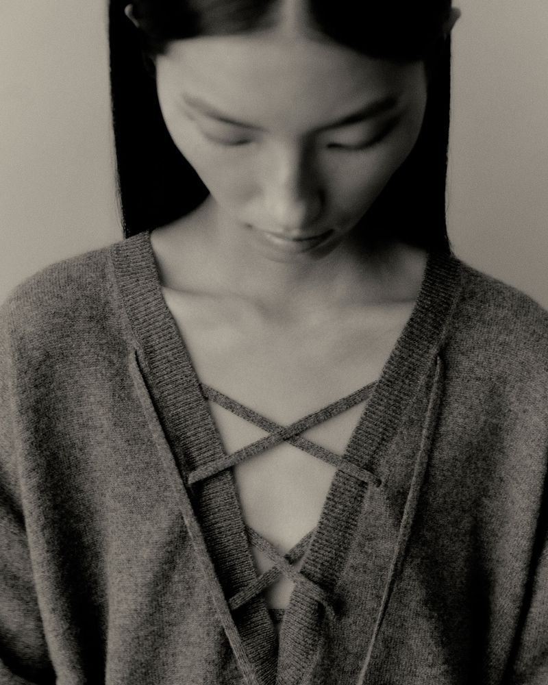 Close-up shot of an Asian woman in a grey sweater with a criss-cross tie detail, eyes downcast, in a neutral indoor setting.