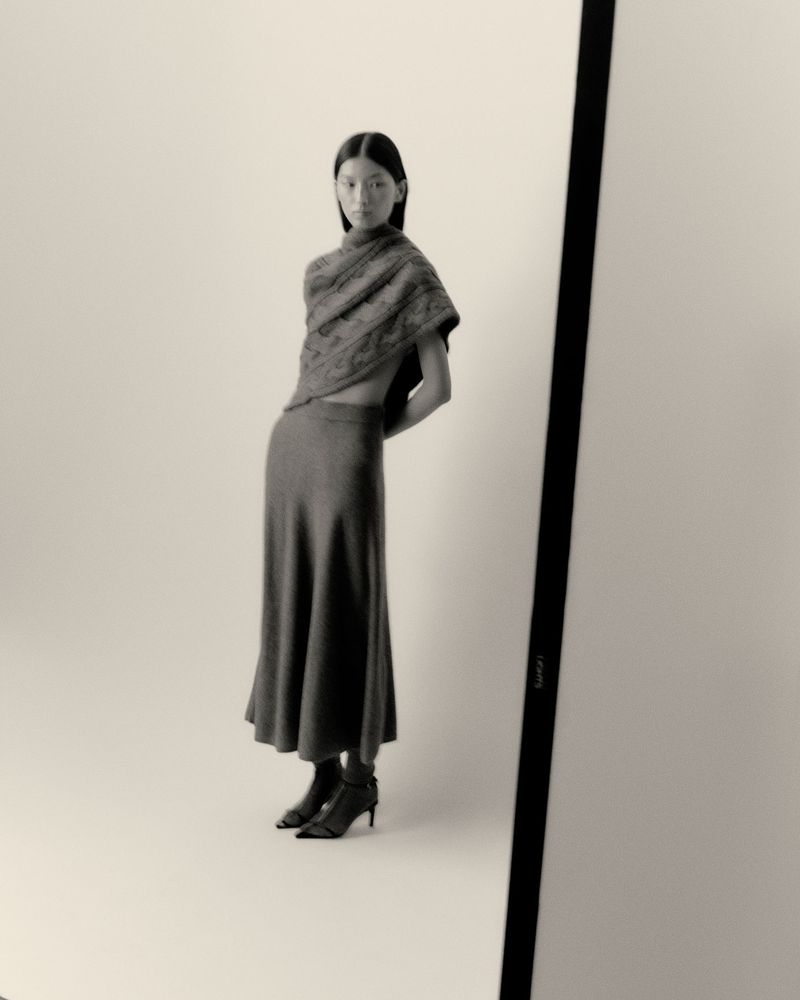 Monochrome image of an Asian woman wearing a knit shawl and skirt, posing in a photo studio with arms behind her back.