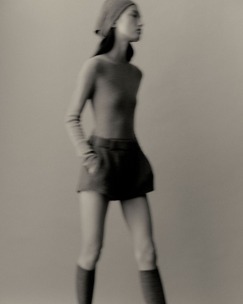 Monochrome fashion portrait of a woman wearing a knit beret, top, shorts and knee socks, standing against a plain background.