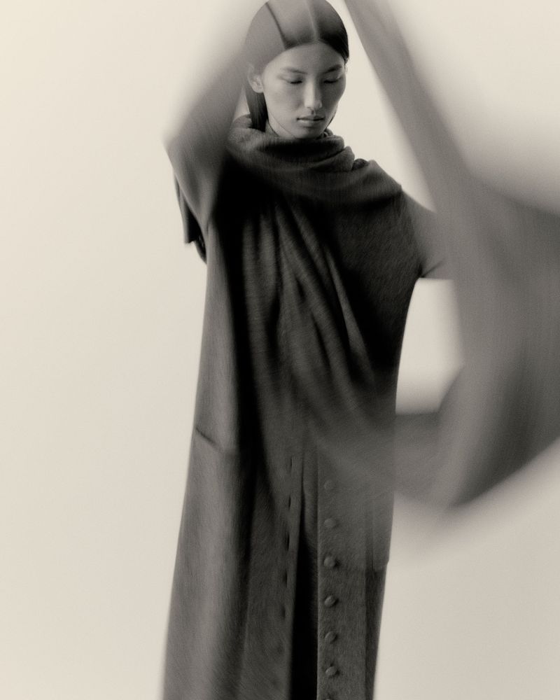 Monochrome studio shot of an Asian woman in a dark shawl-necked vest with button details, soft diffused lighting, and motion blur.