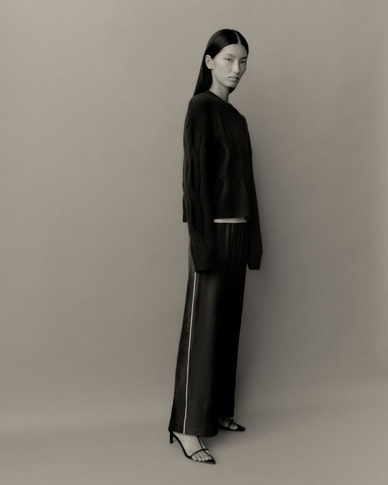 Model wearing a black sweater and dark trousers standing against a neutral grey studio backdrop