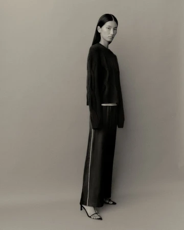 Model wearing a black sweater and dark trousers standing against a neutral grey studio backdrop