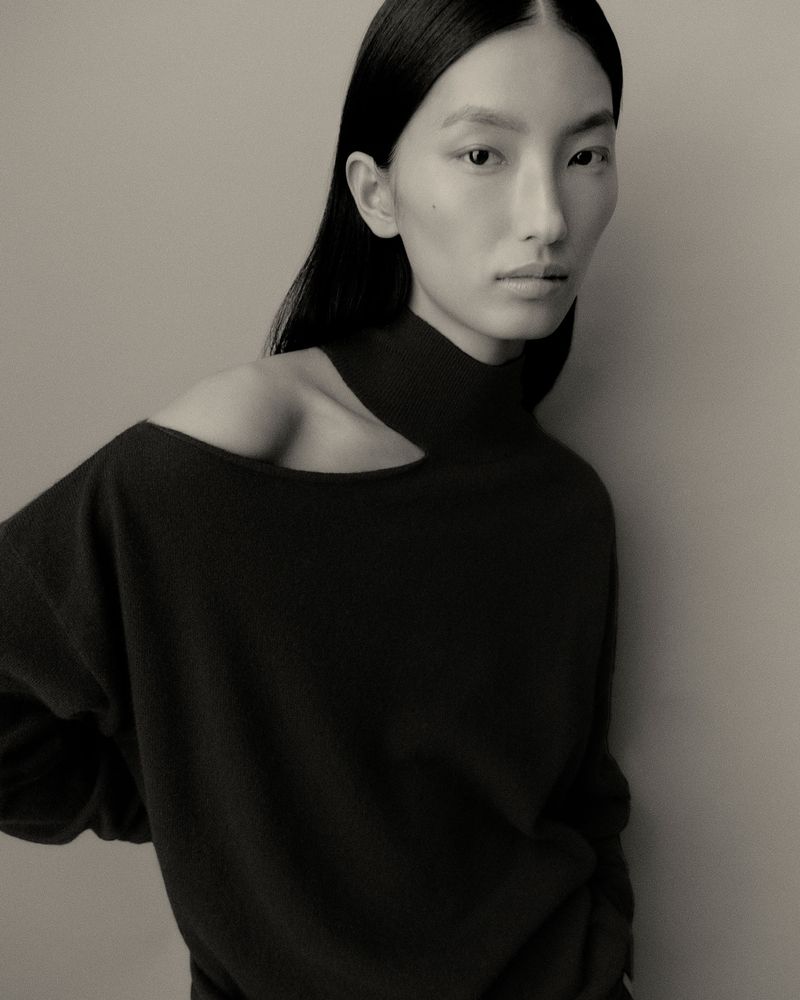 Model with long dark hair wearing a black asymmetrical knit sweater against a plain neutral backdrop