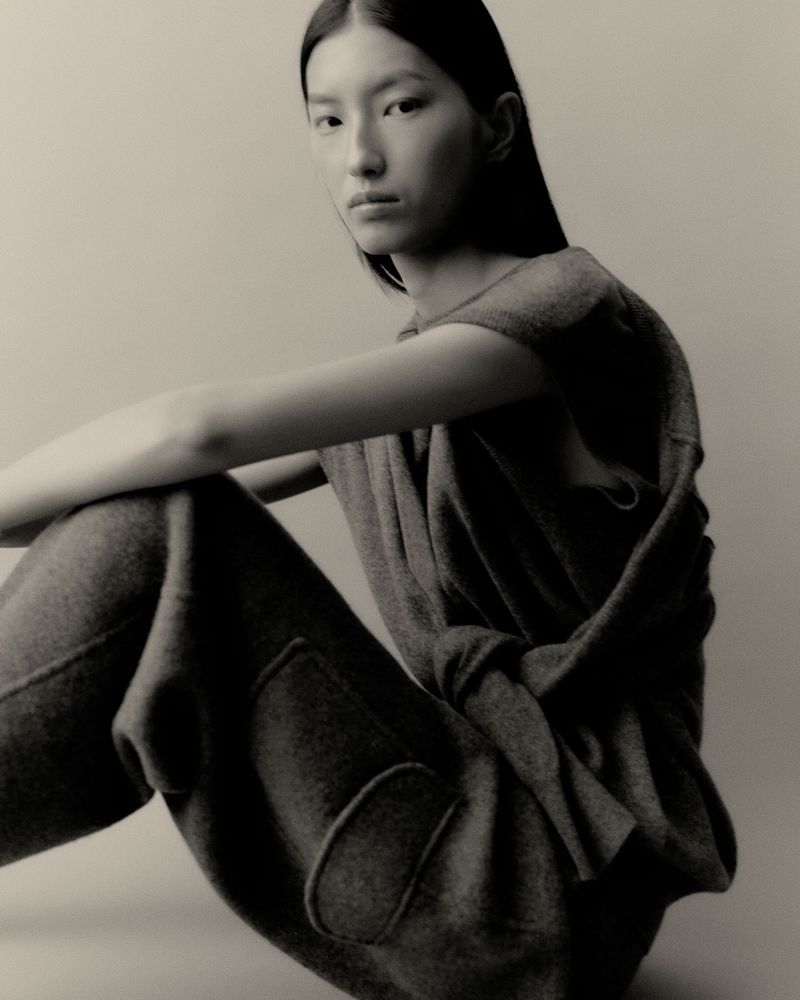 Portrait of a contemplative young Asian woman in a grey knit top and pants seated in a photo studio with neutral lighting.