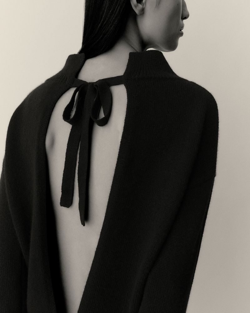 Back view of an adult female fashion model wearing a black knit sweater dress with a tied bow. Cream-colored background. Studio lighting.