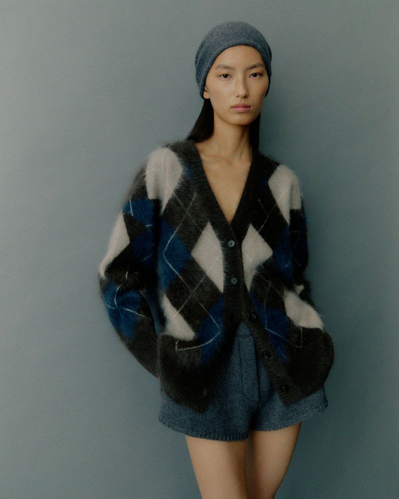 Adult woman in a fuzzy argyle cardigan and shorts poses in a studio with soft lighting. She wears a matching beanie.