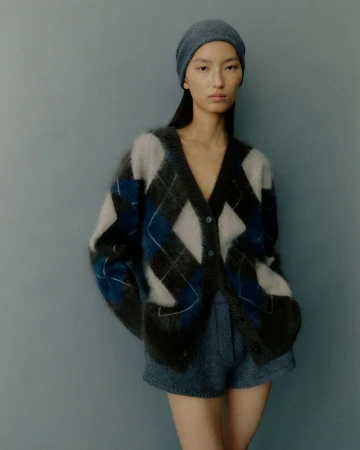 Adult woman in a fuzzy argyle cardigan and shorts poses in a studio with soft lighting. She wears a matching beanie.