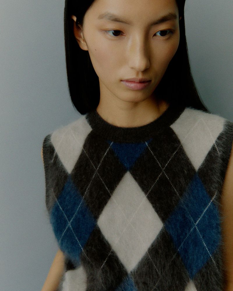 Close-up of an adult woman with fair skin and long dark hair, wearing an argyle sweater vest against a light blue background.