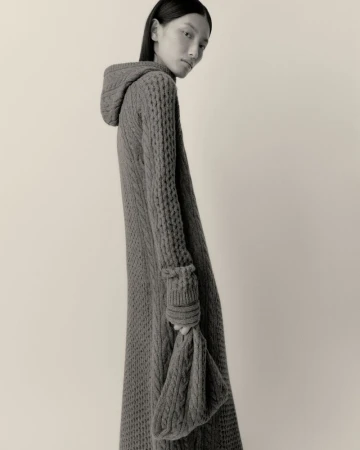 Model Xinyue Guo wearing a long grey hooded cable-knit dress and holding a matching knit bag against a studio backdrop.