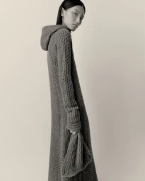 Model Xinyue Guo wearing a long grey hooded cable-knit dress and holding a matching knit bag against a studio backdrop.