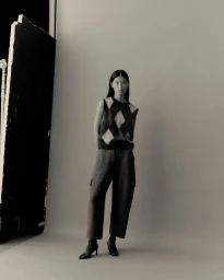 Model Xinyue Guo stands wearing an argyle knit vest, cropped trousers, and pointed heeled boots against a grey studio backdrop.