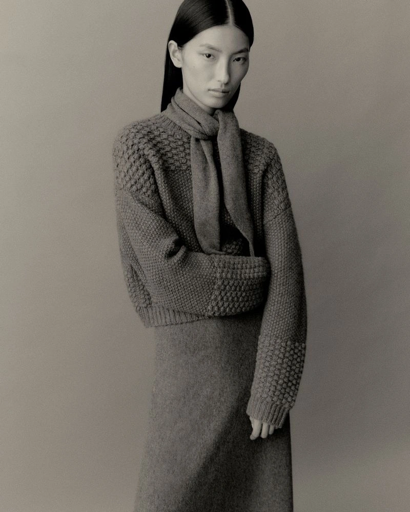 Model wearing a textured knit sweater and matching skirt with a scarf against a neutral studio backdrop