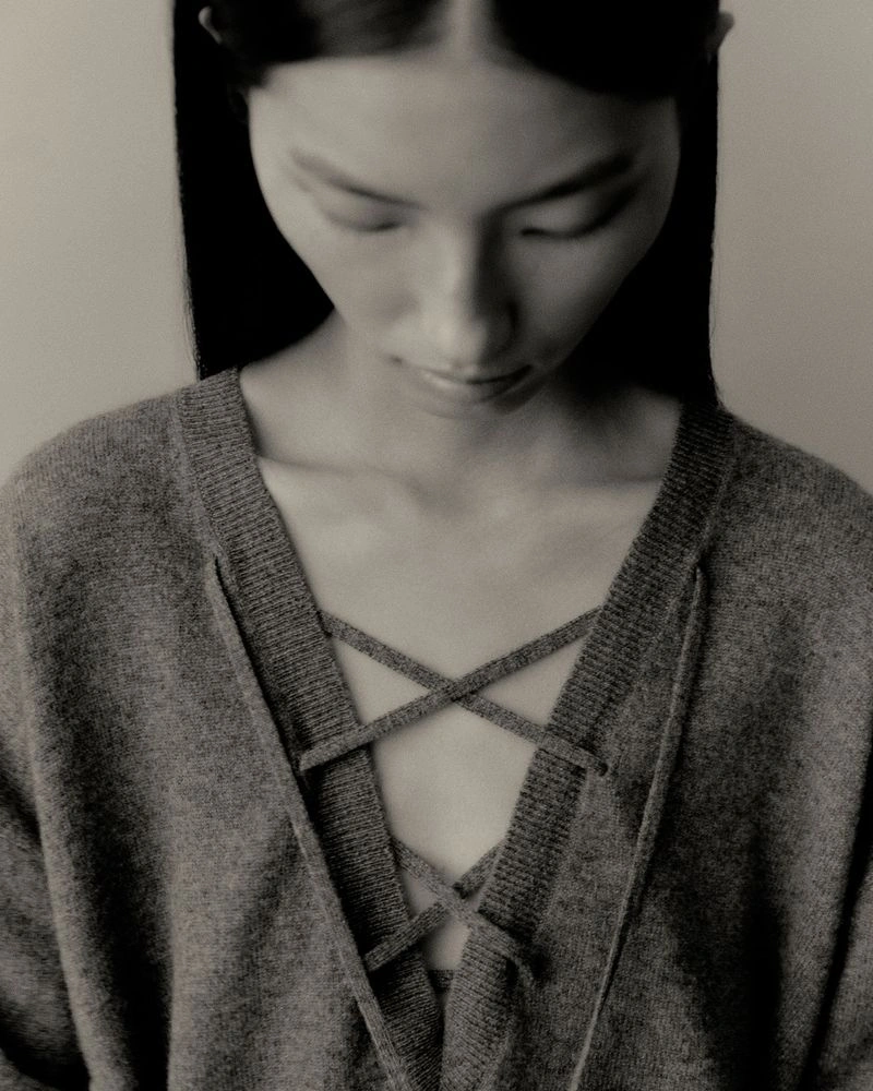 Close-up portrait of a model wearing a grey knit sweater with lace-up neckline details against a neutral background.