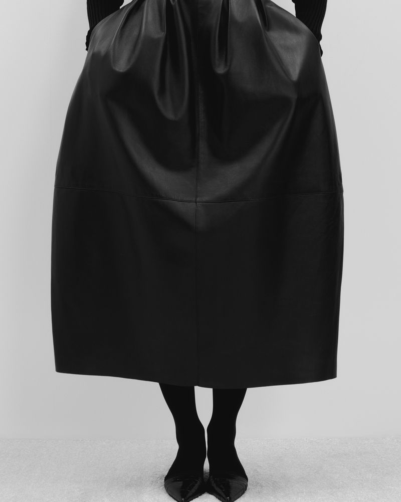 Person wears a black leather midi skirt with a gathered waist, paired with black pointed heels and black tights on a plain background.