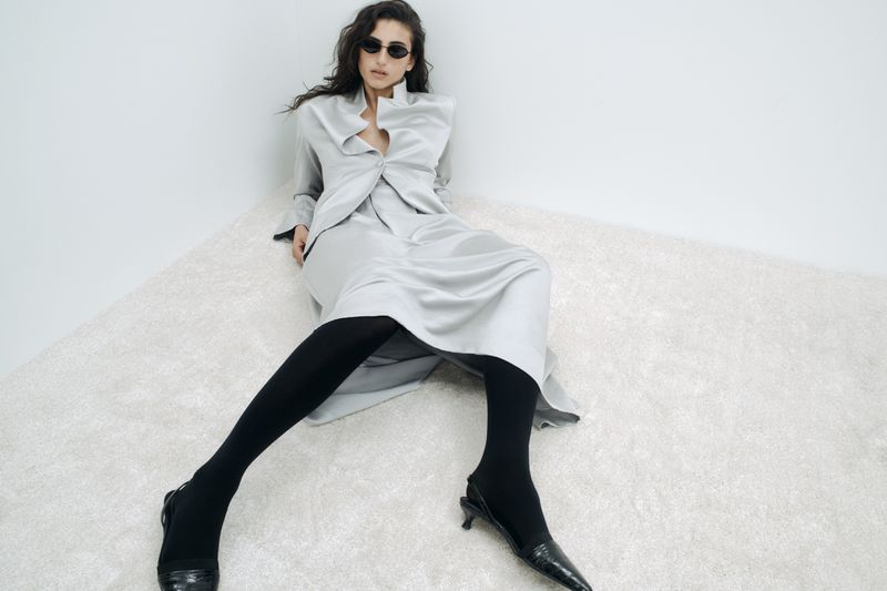 Fashion shot of a woman in a grey blazer and skirt, posing on a shag carpet in front of a white wall, wearing sunglasses.