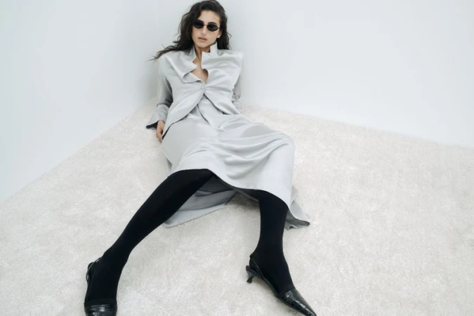 Fashion shot of a woman in a grey blazer and skirt, posing on a shag carpet in front of a white wall, wearing sunglasses.