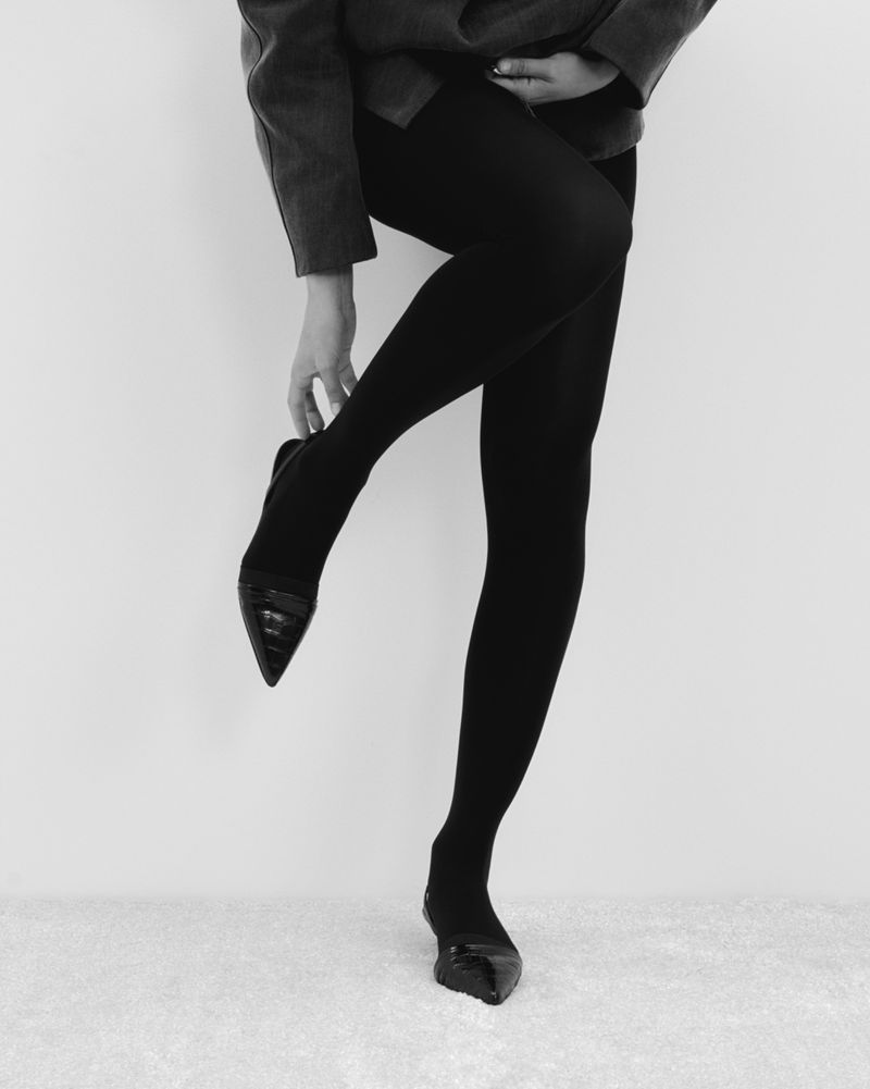 Black and white studio shot of a woman wearing black tights and pointy high heels, holding one foot, and wearing a jacket.