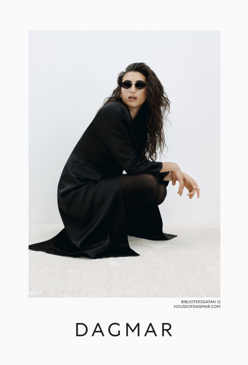 A woman in a dark outfit and sunglasses poses in a crouched position indoors against a light background, showcasing high-fashion elegance.