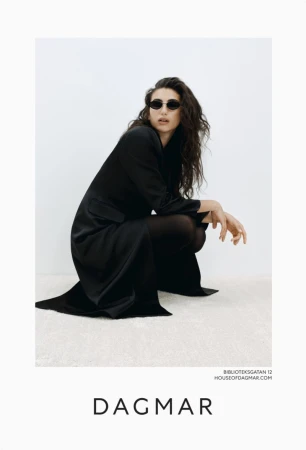 A woman in a dark outfit and sunglasses poses in a crouched position indoors against a light background, showcasing high-fashion elegance.